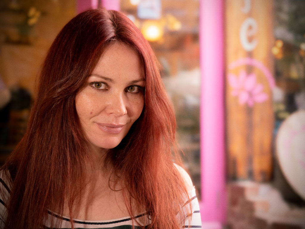 Portrait of a woman with long red hair smiling at the camera, in the background out of focus shopfront