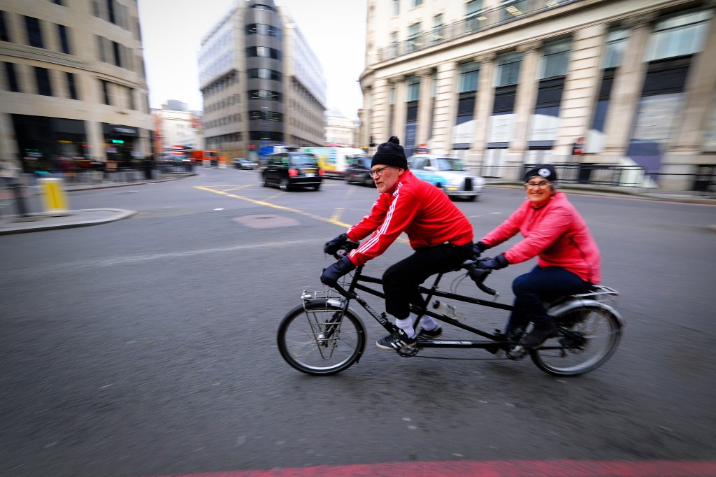 Panning shot of tandem cyclists in a busy London street, dressed in matching black trousers and red raincoats. Shot on Fujifilm X-H2S