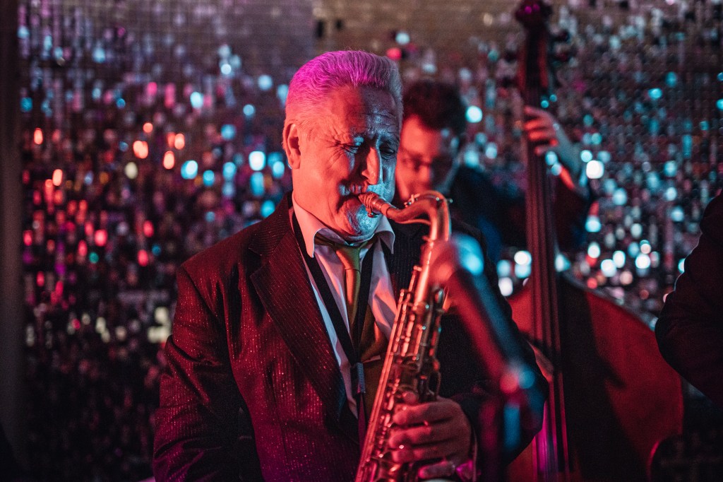Man playing saxophone , another person in the background plays double bass, purple and blue stage lights, silvery glittering out of focus background.