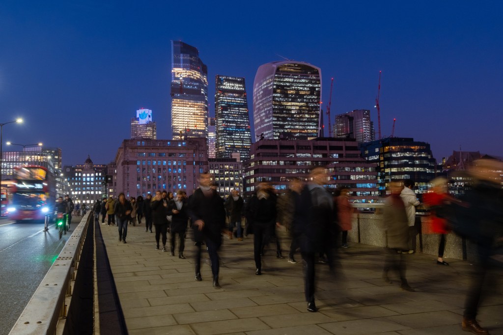Long exposure, motion blurred image of people walking on London Bridge at night. Skyscrapers in focus in the background set against the night sky.