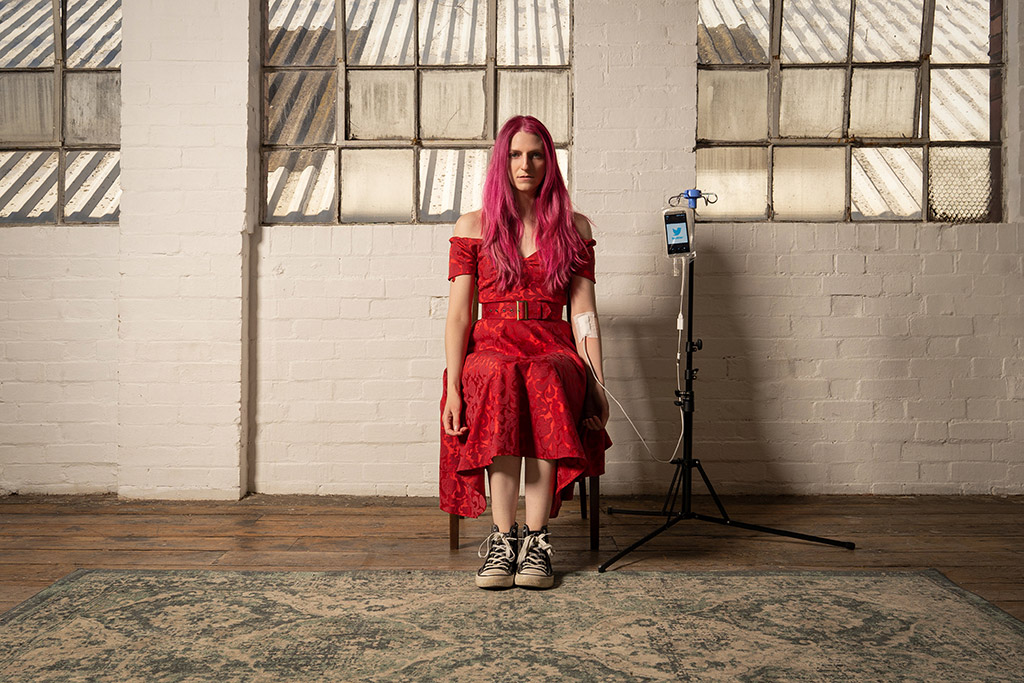 woman with pink long hair dressed in red sat on a chair set up with phone in drip