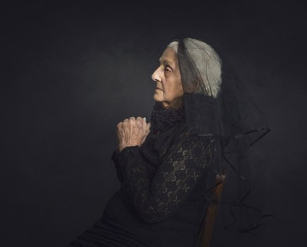 older woman in black dress and veil sat against black background
