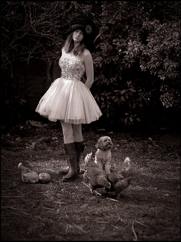Evie Parry with Hens. Girl in a fancy dress and hat standing in a garden, a dog a chickens at her feet.