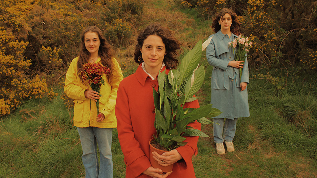 girl in red coat holding house plant with two girls behind holding bouquets