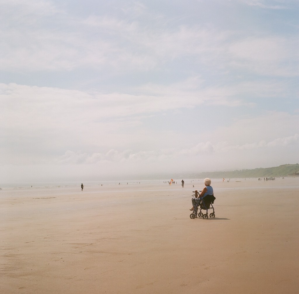 Elderly person in a wheelchair on an empty sandy beach, looking out to the see