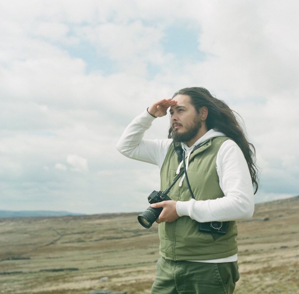 Man with long hair and camera looking into the distance shielding his eyes with his hand