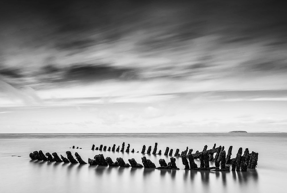 Lightroom vs Photoshop. Final image an old wooden shipwreck sticks out from the sand in Nornen beach near Burnham-on-Sea in Somerset after photoshop black and white editing. 