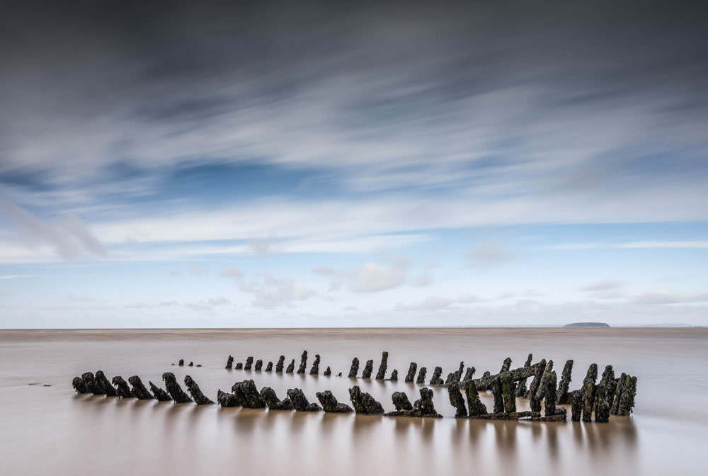 Long exposure shot of blue sky with blurred clouds, the spine of an old wooden shipwreck sticks out from the sand in Nornen beach near Burnham-on-Sea in Somerset. 