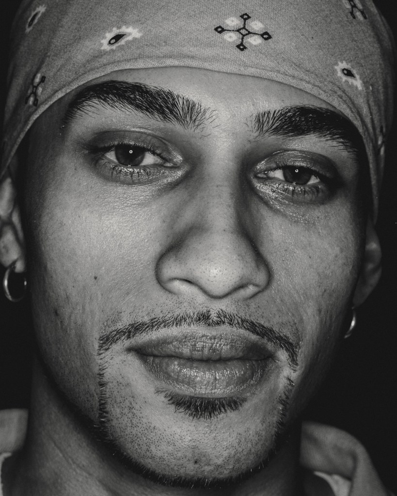 Close up black and white analogue portrait shot of a man with thin mustache, and beard. Two silver earrings and bandana on his head, smiling slightly at the camera