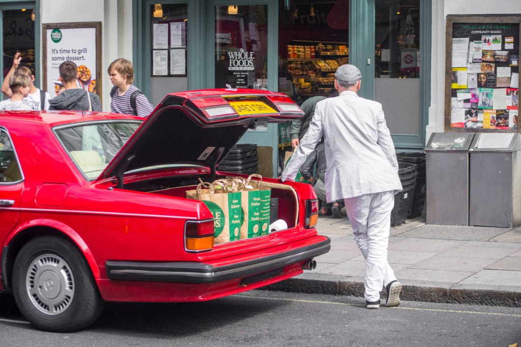 Man in a white suit and grey flatcap leaning against the back of a red car. trunk open shopping bags lined up inside