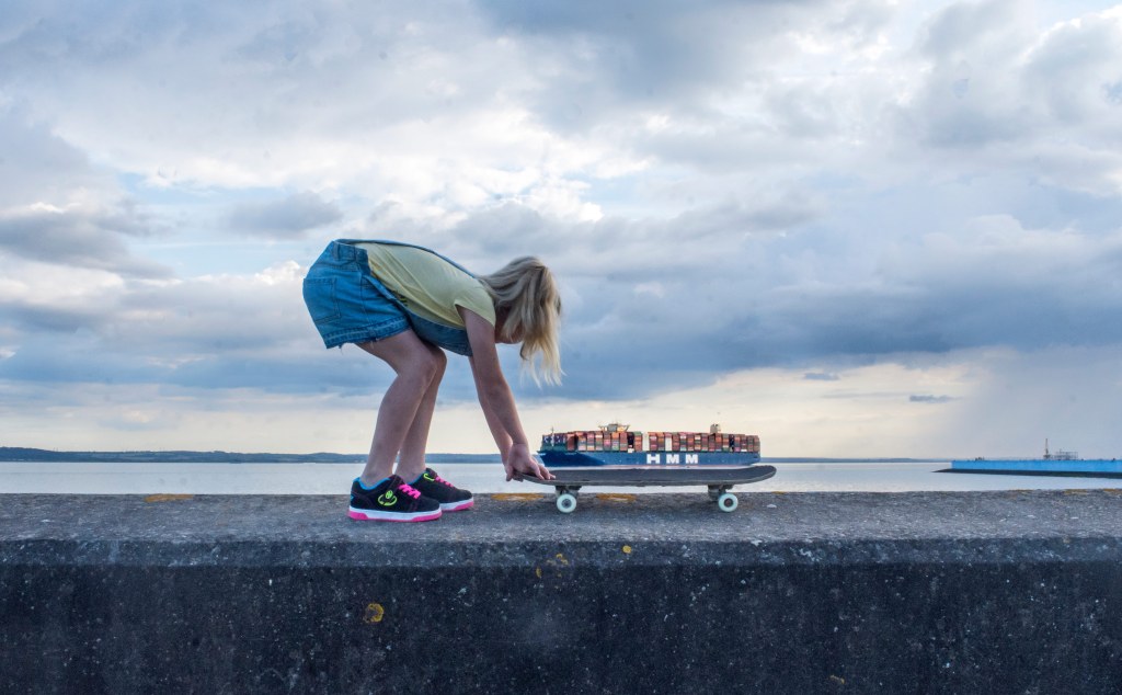 A girl pulling a skateboard that is aligned with a container ship in a perspective that looks like the ship is hauled on top of the kateboard.