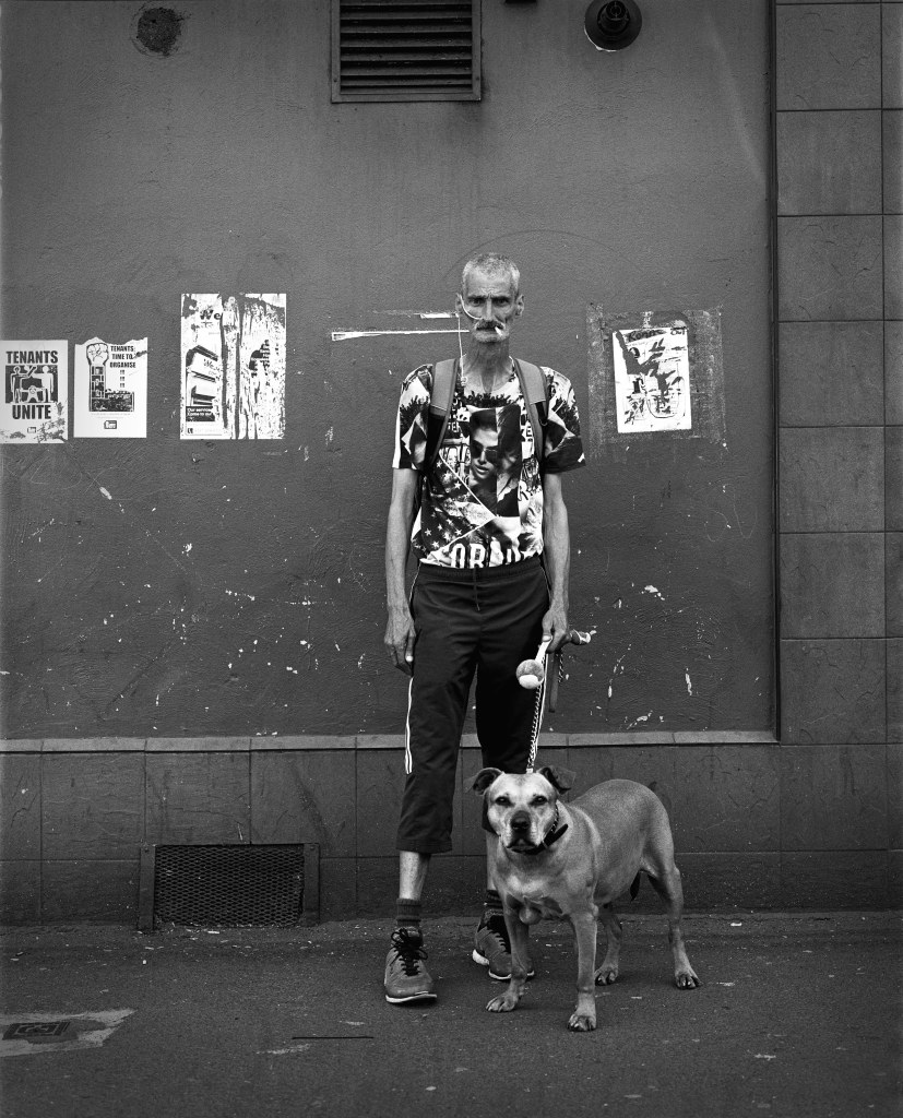 full body portrait of a tall thin older man, with his Staffordshire Terrier. Set againt a wall with old white posters , he stands in a gap of the posters with cigarette in his mouth, a medical tube leading from his nose down to the edge of his shirt.