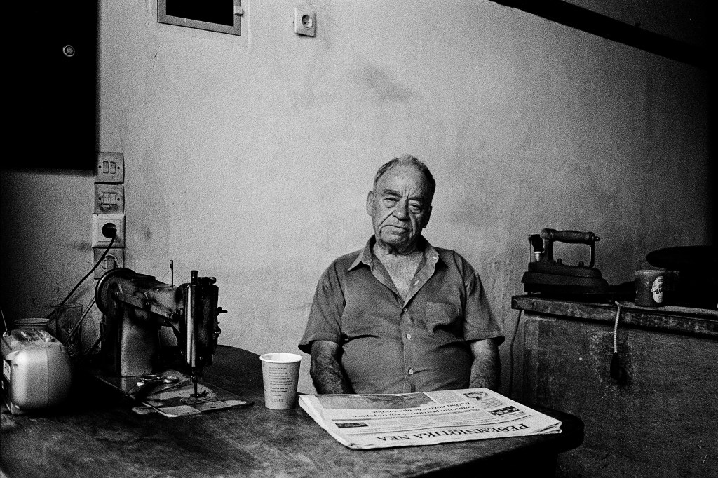 portrait of an older man sitting at sawing machine's table, arms in his lap, a newspaper and a paper cup in front of him. His posture and face relaxed. 