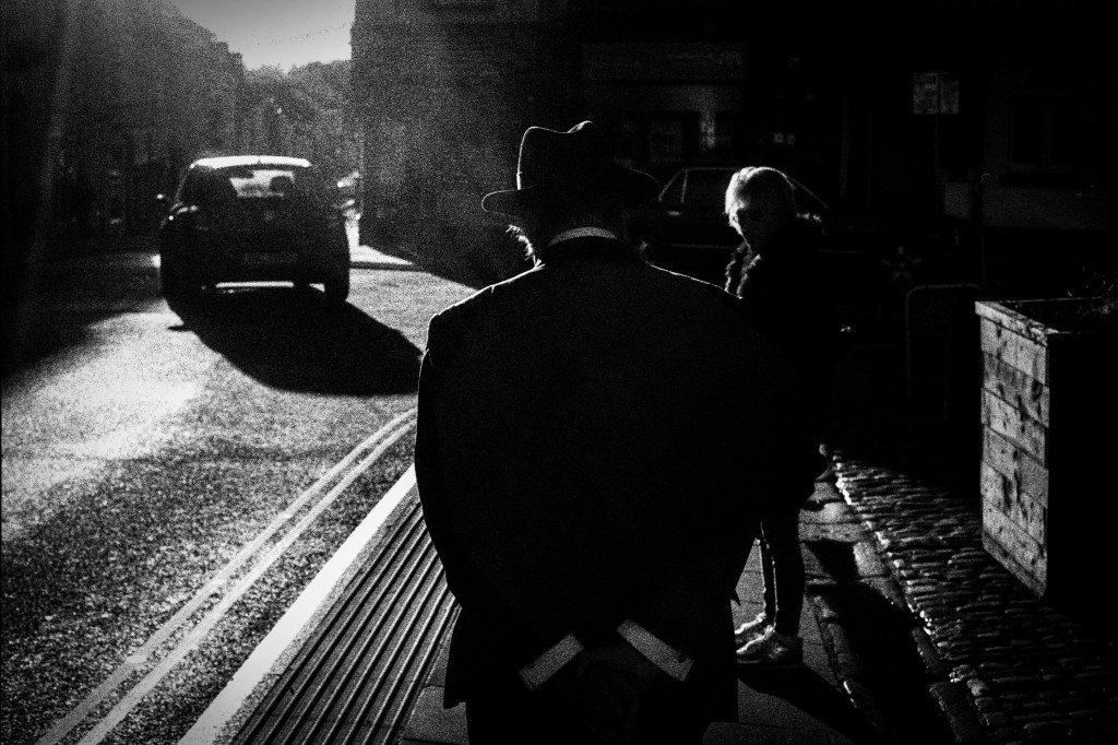 High contrast image of a man walking down the street. Wearing a classic hat, suit and white shirt we see him from behind hands resting on his back. A car passing on the left and another person standing on the right.