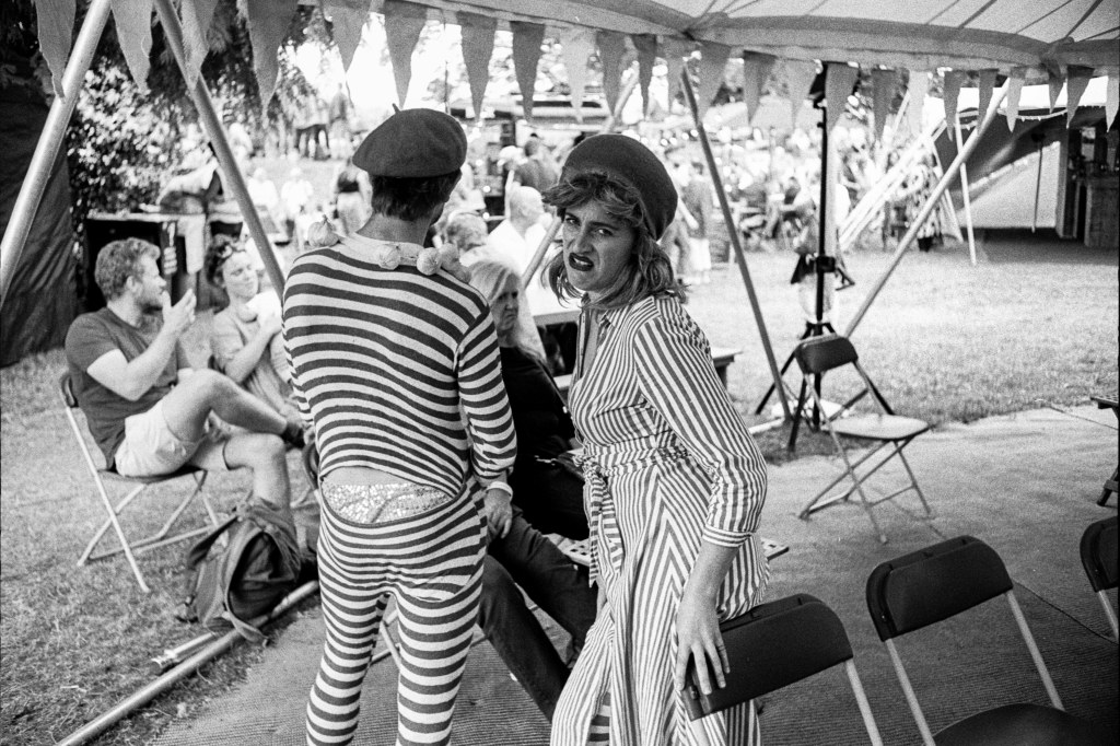 A man and a woman in stripy costume and french hat standing under a tent with banters on its side. In the background other tents and people in what appears to be a fair or festival.