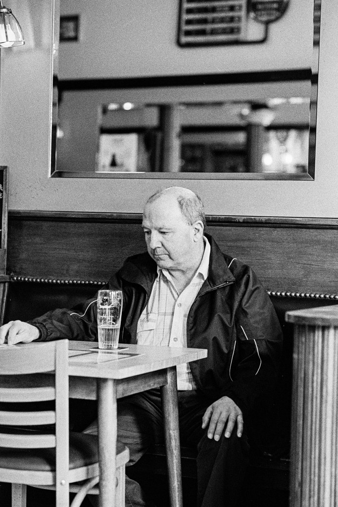 Black and white portrait of a man sitting by the wall in a pub. One hand on top of the table other on his knee, staring at a pint glass in front of him on the table.