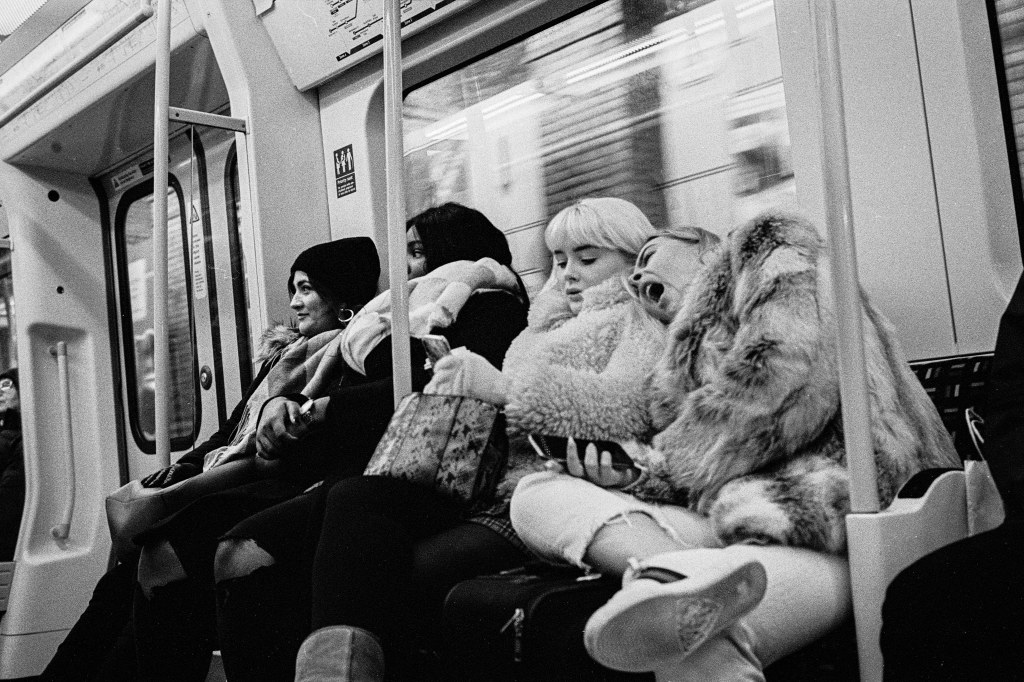 Black and white portrait of four girls in big coats sitting on the undergound. One leans on the shoulder of the other yawning.