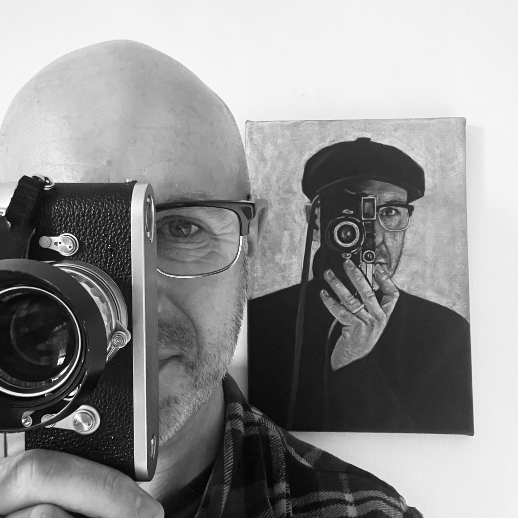 Portrait of David COllier with a camera in front of his face, in the background on the wall an image of him in the same pose and camera in a hat.
