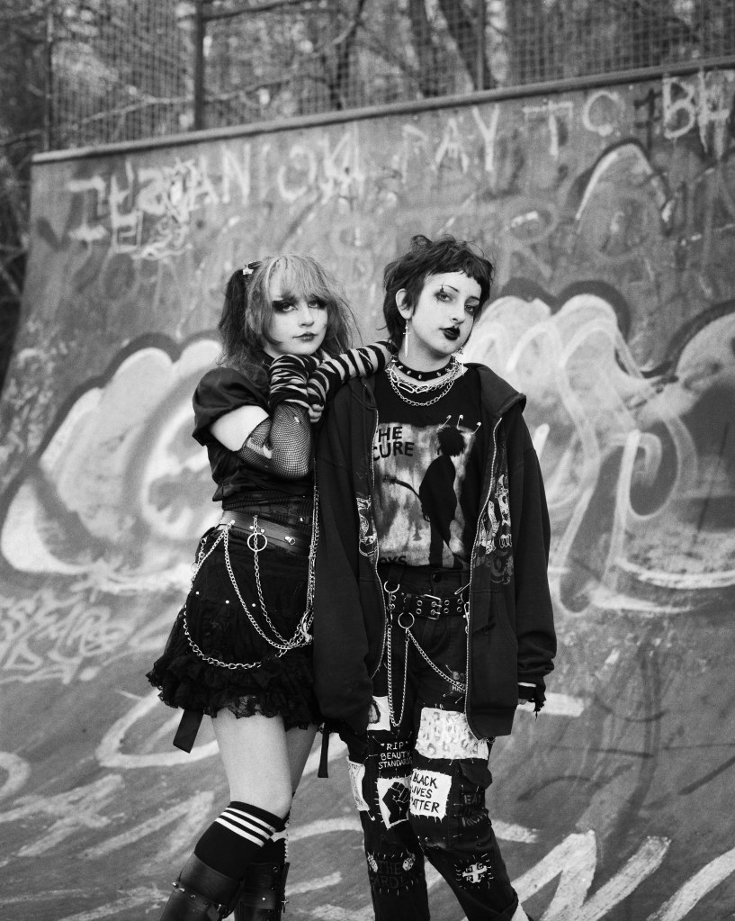 Analogue black and white shot of two teenagers dressed in emo punk clothes. The girl leaning on the shoulder of the boy arms crossed and resting her head on top.