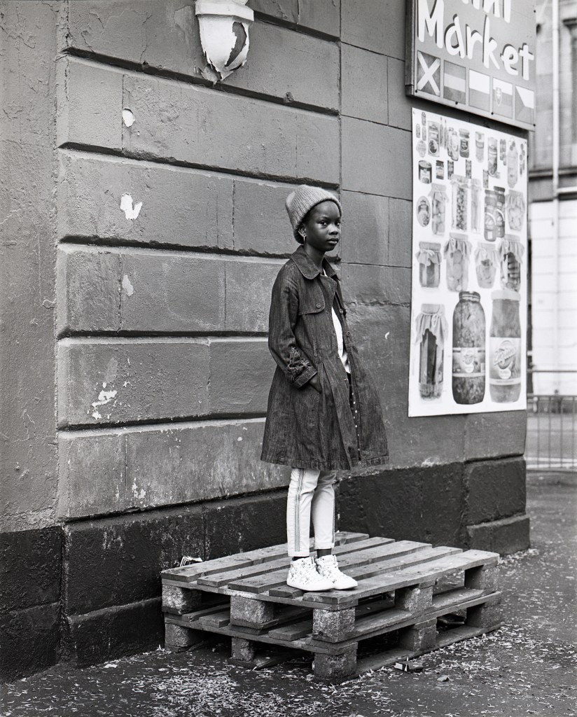 black and white analogue shot. A young black girl in coat and hat, hands in her pockets standing on the corner of stacked pallets in front of a grocery store. 
