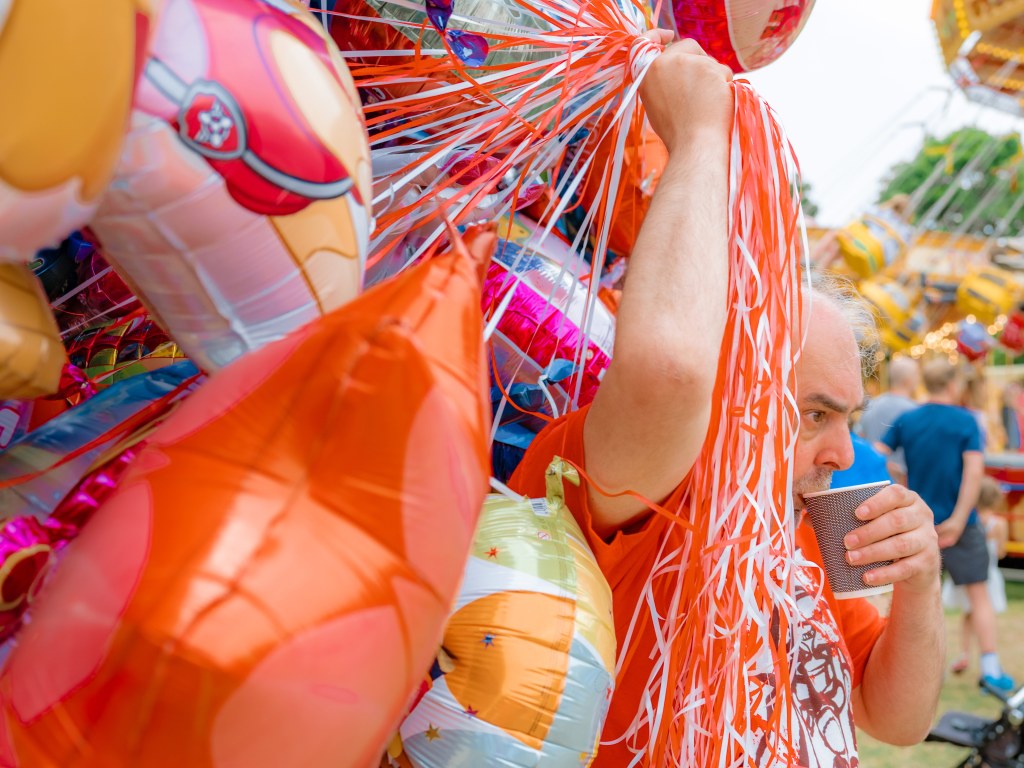 Man at a fair sipping from a paper cup holding dozens of balloons that cover most of the frame