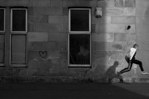 simon murphy black and white analogue photograph of a boy jumping, a streak of light illuminating him ,his shadow cast on the building next to him