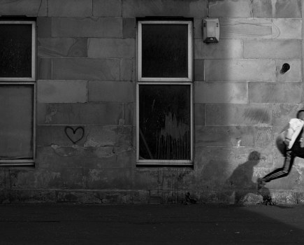 simon murphy black and white analogue photograph of a boy jumping, a streak of light illuminating him ,his shadow cast on the building next to him