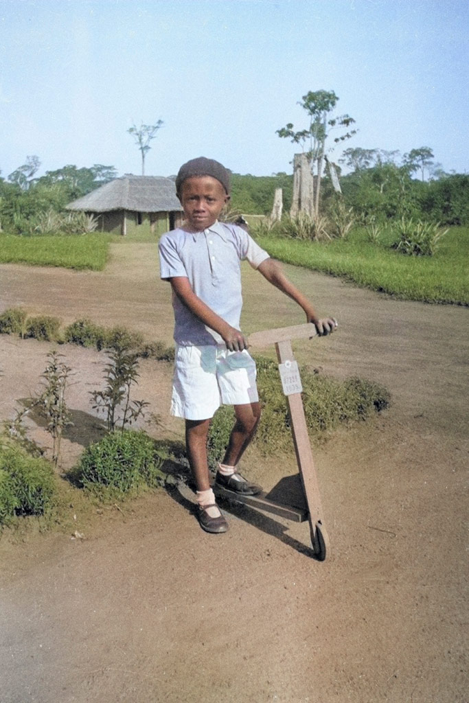 Young Black boy poses on a wooden scooter, in a green rural setting. Photoshop AI colouring example