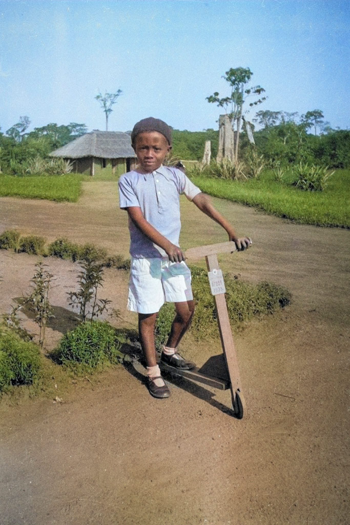 Young Black boy poses on a wooden scooter, in a green rural setting. Photoshop AI colouring example