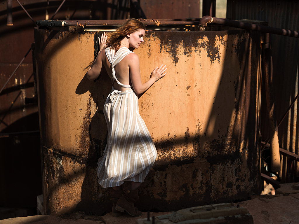 model in white dress posing against orange wall with strong light against her and shadows surrounding