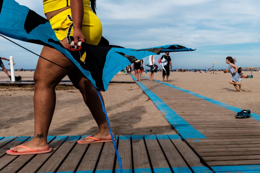 A woman in a yellow outfit on the beach, carrying a blue kite. Blue lines on the wooden path connect the subject and create strong leading lines.