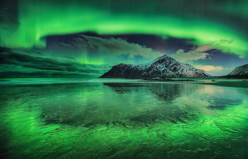 A stunning photograph of a vivid aurora over Skagsanden beach, Lofoten Islands, Norway. The mountain in the background is Hustinden, which the aurora appears to encircle. 