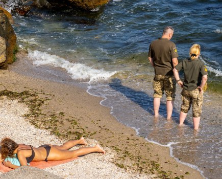 A man and a woman in Ukrainian military outfit, with trousers rolled up standing in the black sea, behind them on the beach a woman sunbathing.