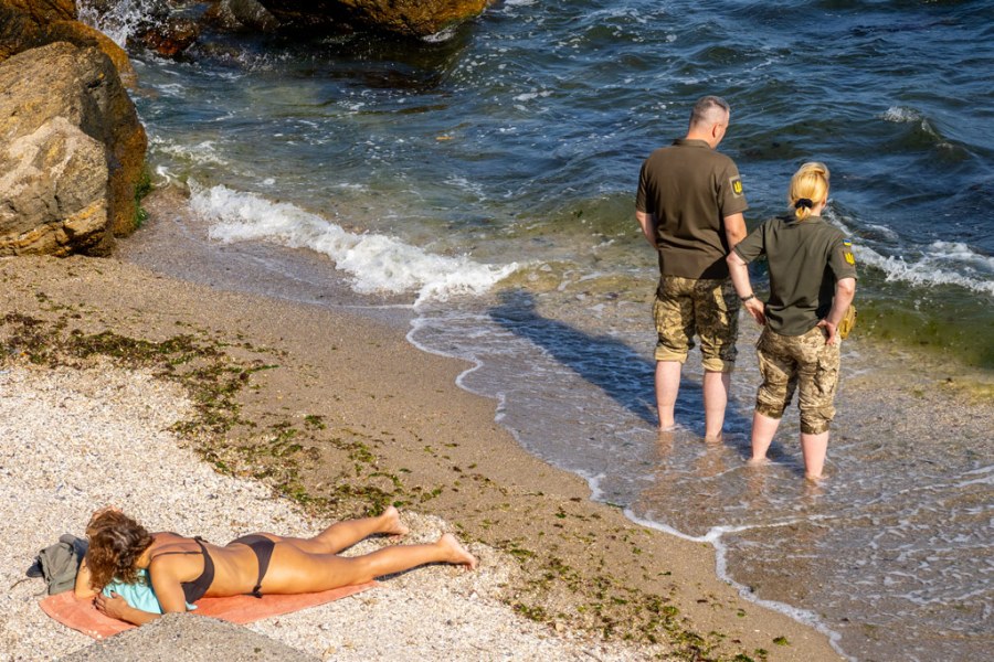 A man and a woman in Ukrainian military outfit, with trousers rolled up standing in the black sea, behind them on the beach a woman sunbathing.