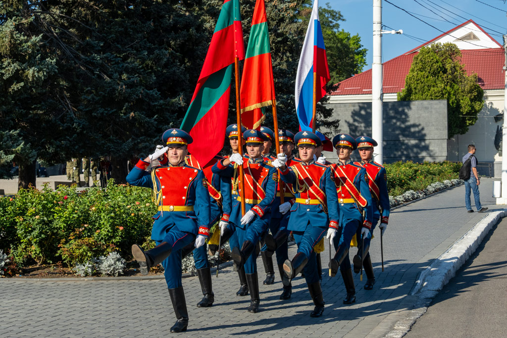 A group of men wearing red and blue military uniform carrying flags march on the street
