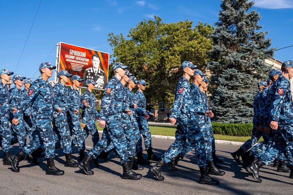 A group of young troops marching on the street in blue military uniform, a billboard in the background features an older man in military uniform and many militray badges