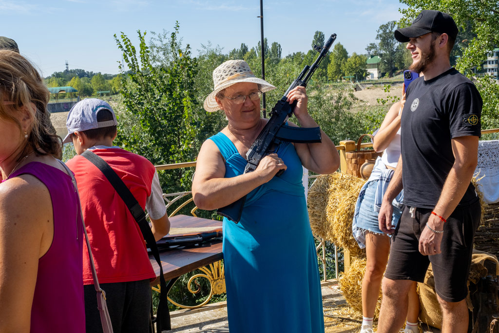 Older woman in a long blue dress and white straw hat poses with a large machine gun, 