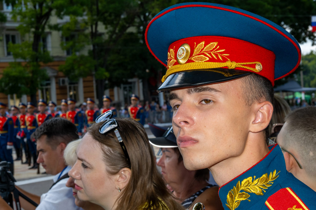 Portrait of a young man standing in the crowd wearing the red and blue military uniform and hat of the Honor Guard of the Pridnestrovian Moldavian Republic