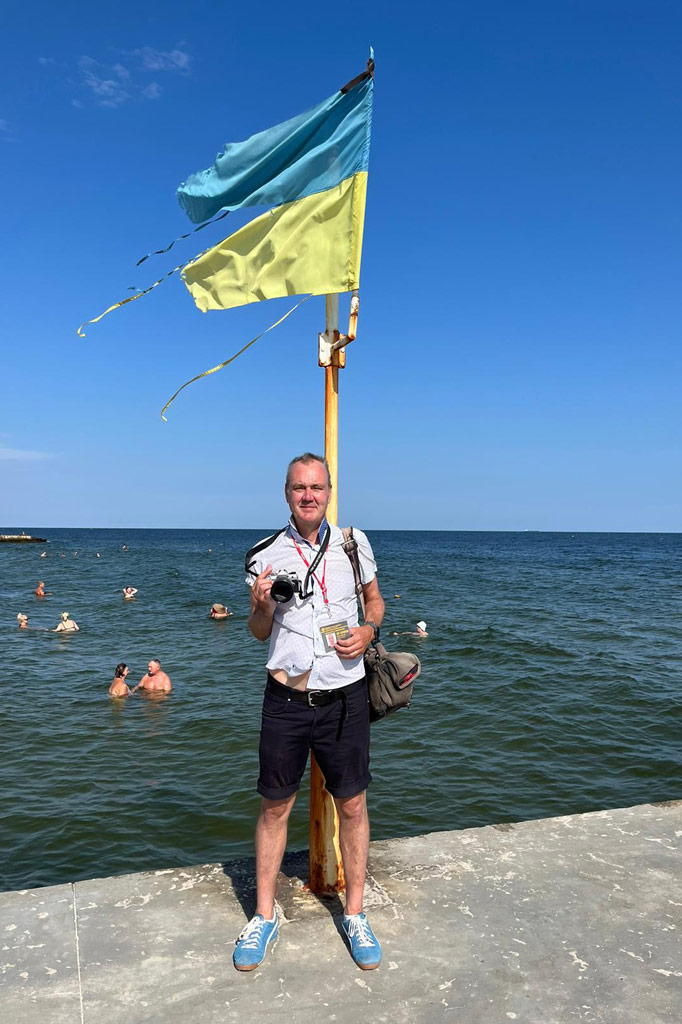 Peter Dench posing with his camera in front of the Ukrainian flag in Odessa, by the Black Sea