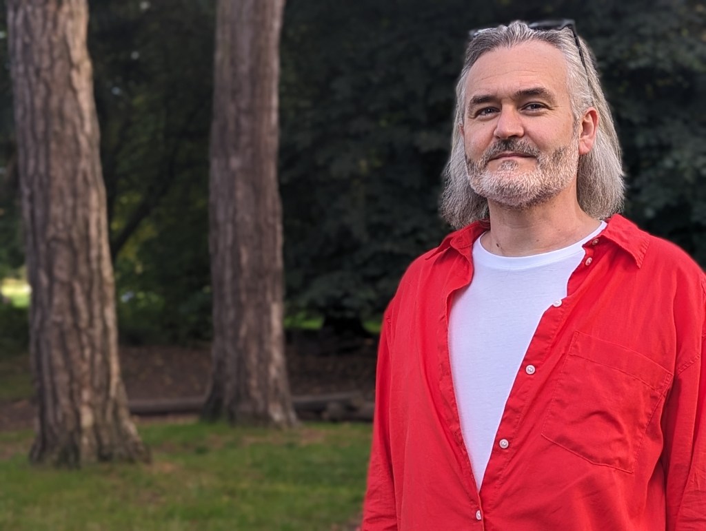 Google Pixel 7 Pro portrait sample image, man with white shoulder length hair wearing a red shirt standing in a park, trees and greenery in the background