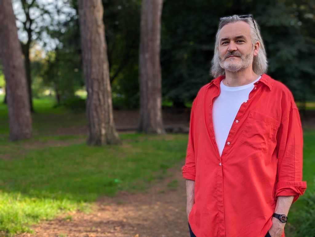 Google Pixel 8 Pro portrait sample image, man with white shoulder length hair wearing a red shirt standing in a park, trees and greenery in the background