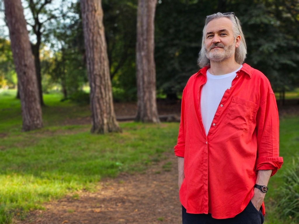 Samsung Galaxy S23 ultra portrait sample image, man with white shoulder length hair wearing a red shirt standing in a park, trees and greenery in the background