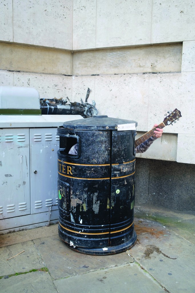 a man sitting inside a black street bin with his guitar's neck sticking out only
