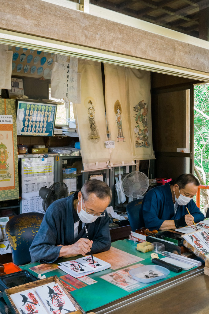 Two Japanese man in blue overcoats doing calligraphy, tradaitional japanese illustrations hang behind them