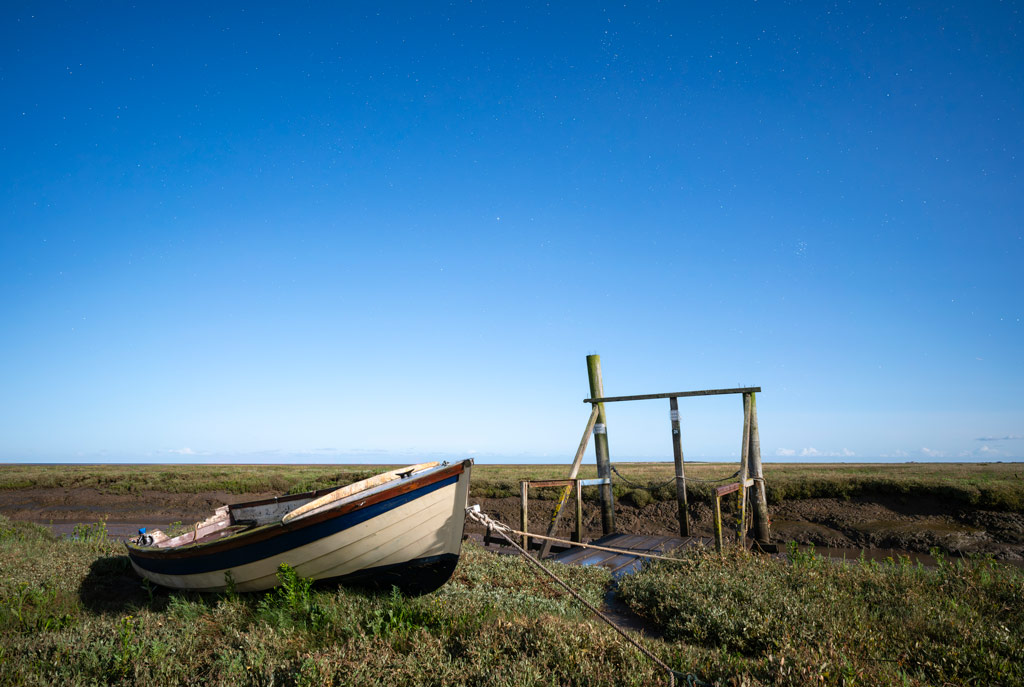 Moonlight landscape with blue starry sky, a small lonely white boat moored on the left side of the image