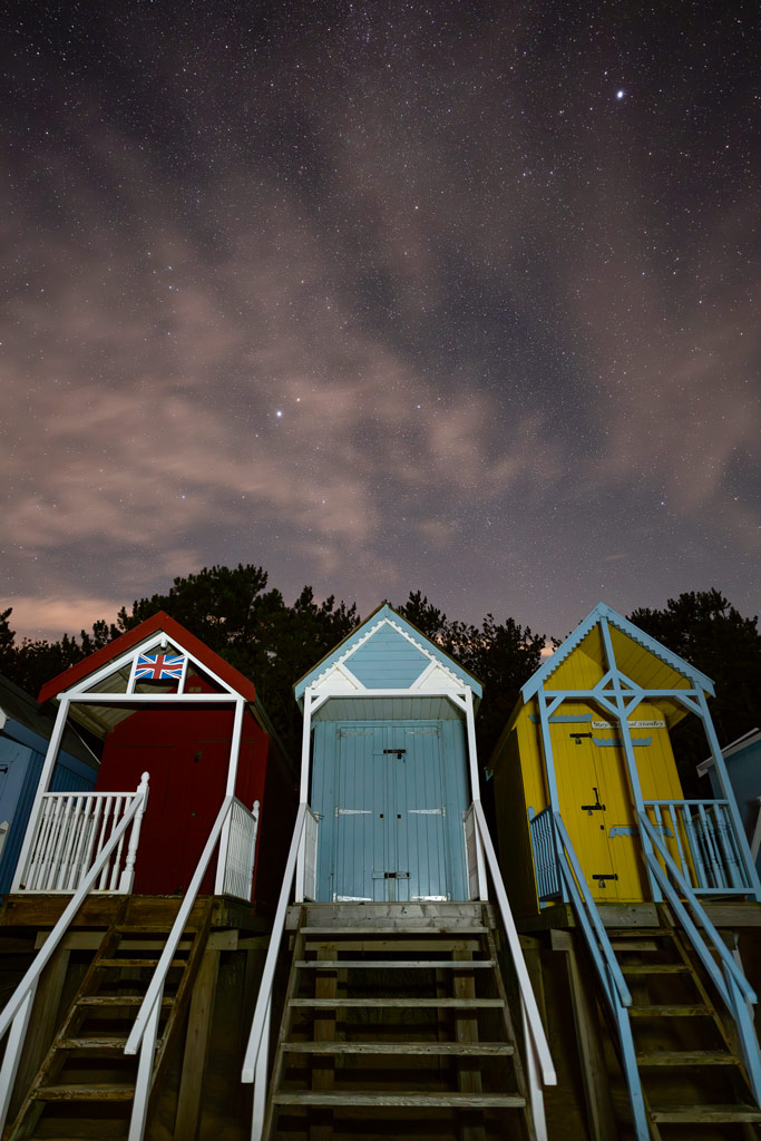 three colourful beach cabins photographed against the a starry and cloudy night sky