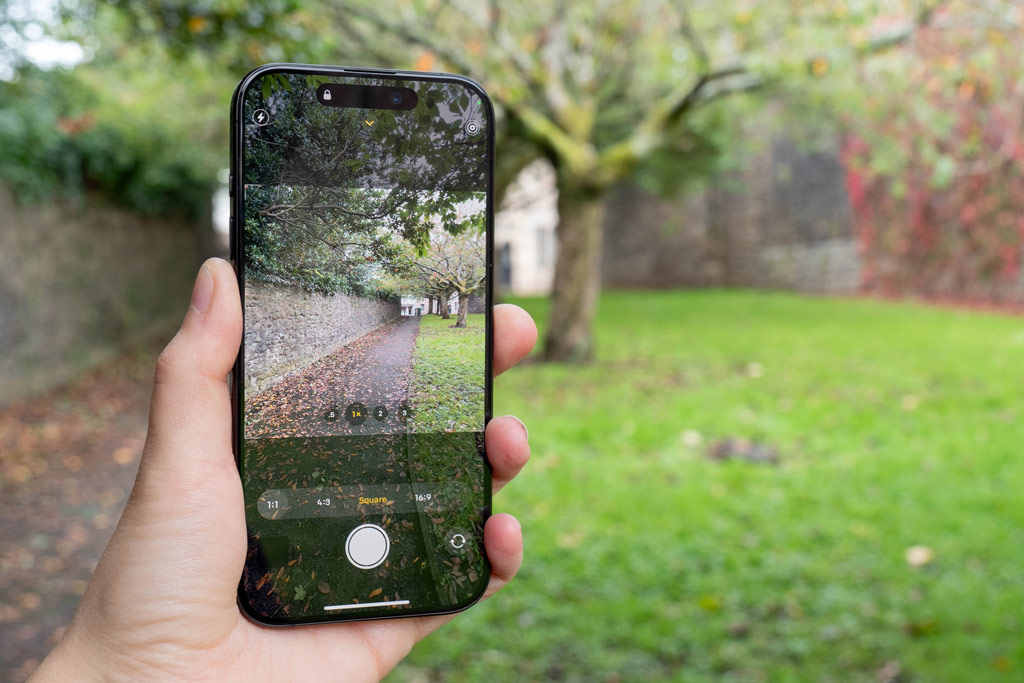 iPhone photography tips. iPhone 15 Pro hold up in hand aspect ratio options in the native camera app, in the background and on the camera screen a path with a tree next to it is showing