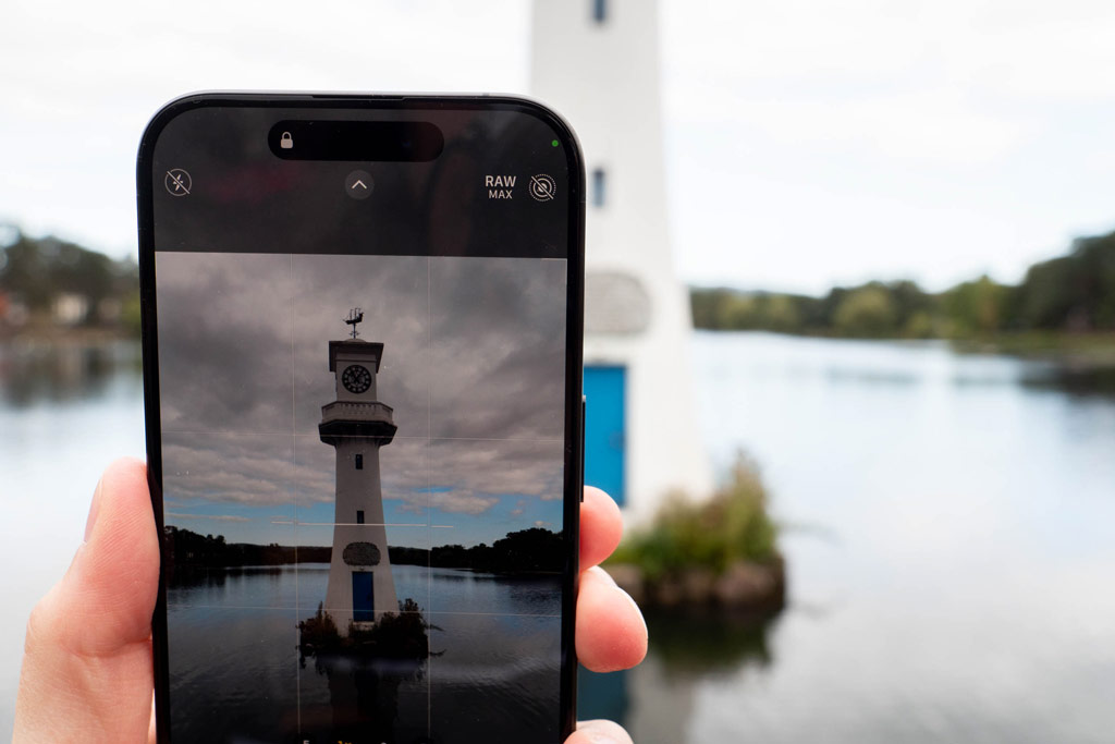iPhone 15 Pro, in hand displaying an image of a lighthouse, in the background the same scene showing
