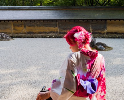 Woman with pink hair wearing a traditional kimono photographed from behind sitting and looking for something in her bag,