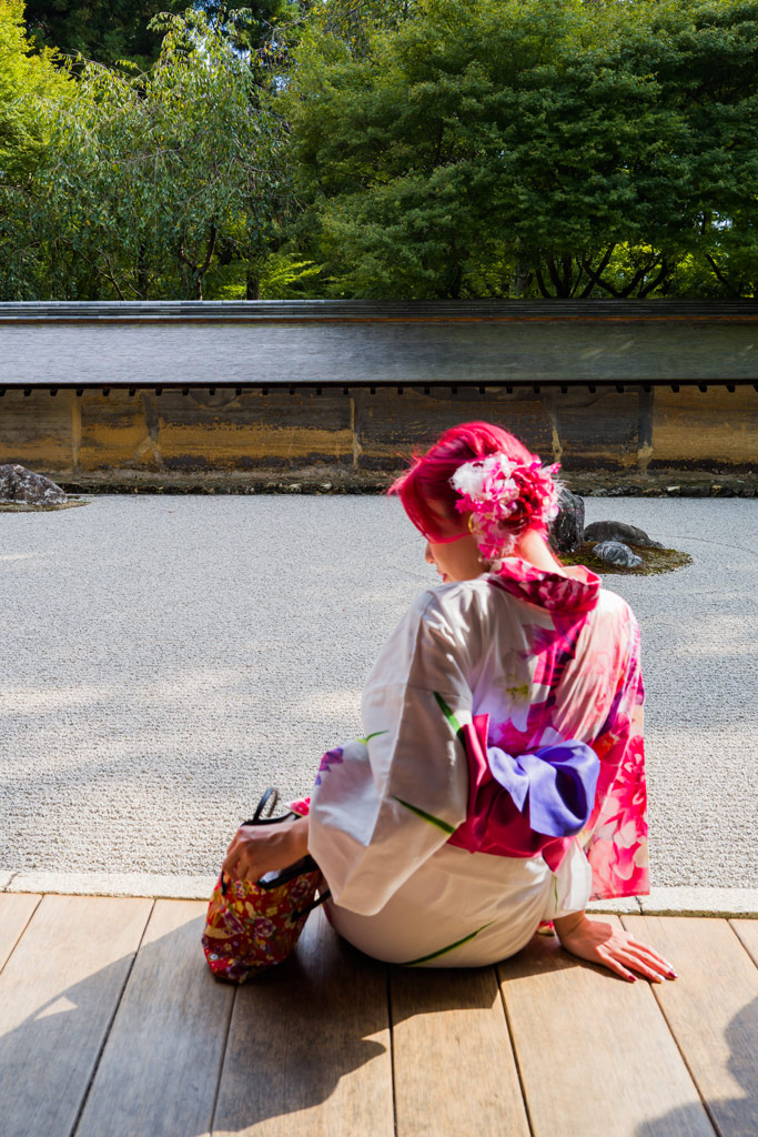 Woman with pink hair wearing a traditional kimono photographed from behind sitting and looking for something in her bag,